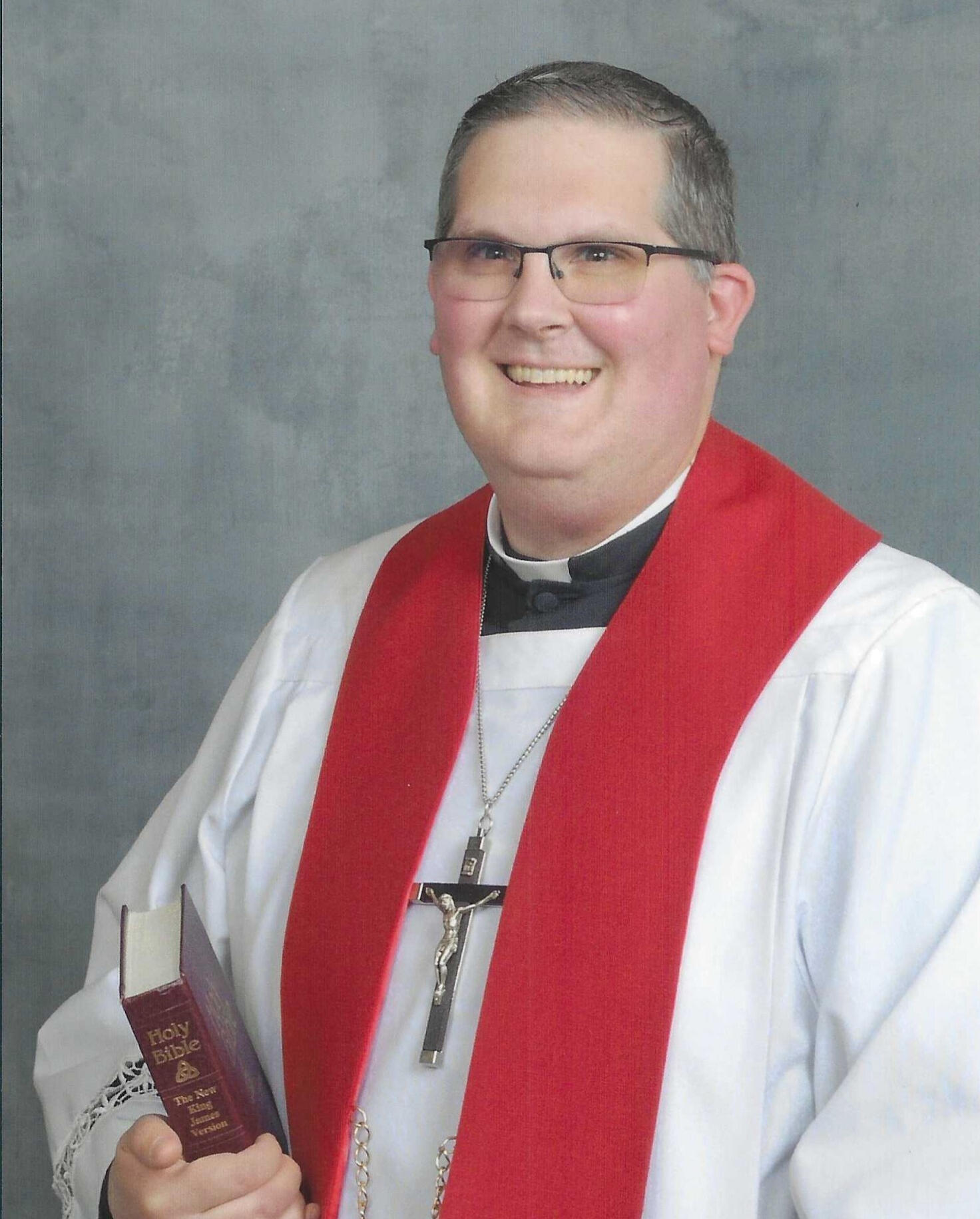 Headshot of Pastor Timothy D Storck of Good Shepherd Lutheran Church in Chesterfield, MI. He is wearing glasses, a large pectoral cross, and a red stole. He is holding a bible and has a cheerful smile.